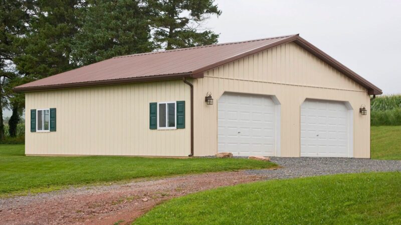 30x32 beige pole barn with brown roof, two garage doors, solid entry door, sliding windows with shutters, and gutters