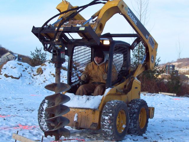 A Timberline Buildings crew member operating a skid steer with a frost-ready auger bit while drilling post holes during winter pole-barn construction in Pennsylvania.
