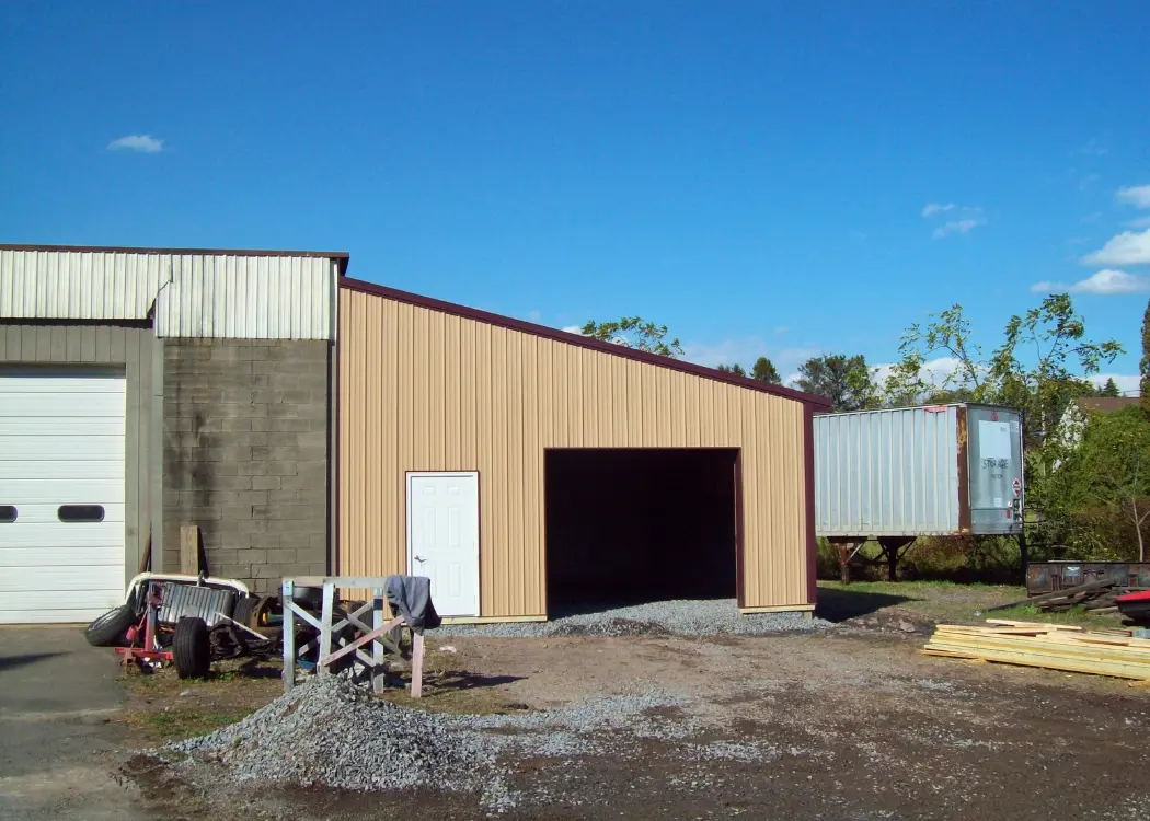 Tan enclosed lean-to attached to a taller building, with a sloping roof, white entry door, and wide vehicle opening.